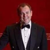 Man in tuxedo smiling against red background.