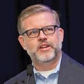 Man speaking into a microphone, wearing glasses.